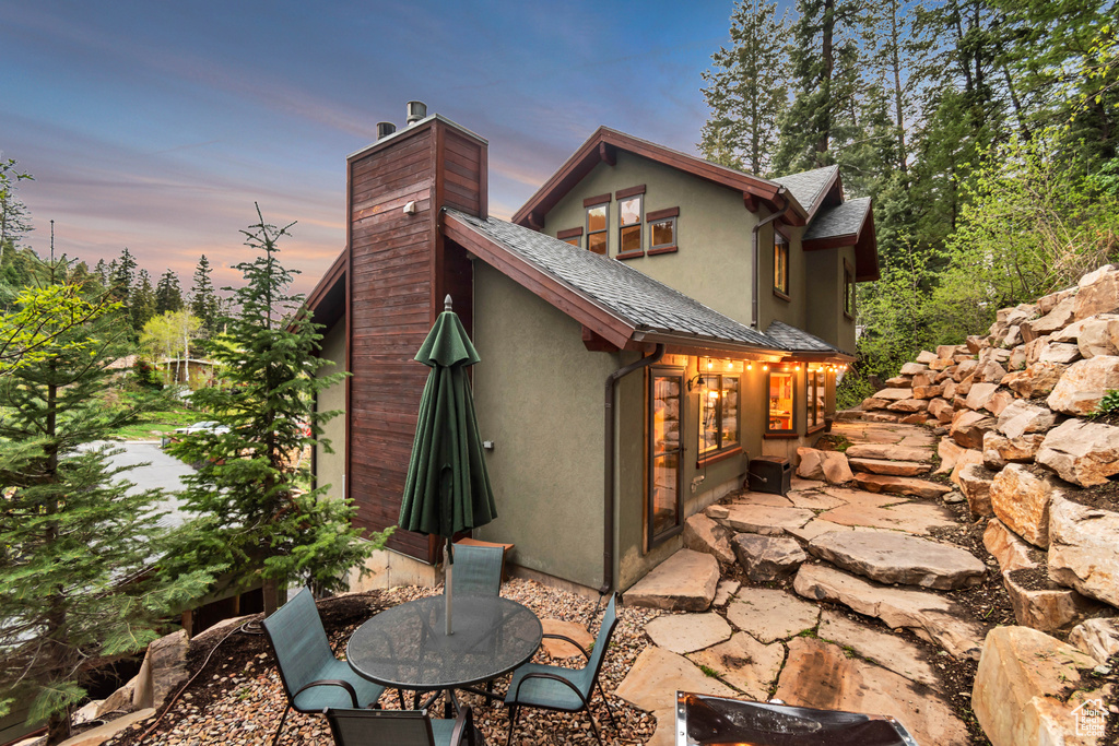 Back of house at dusk with stucco siding, a patio area, a chimney, and a shingled roof