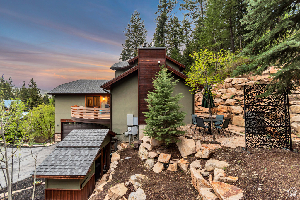 Back of house at dusk with stucco siding, a balcony, a shingled roof, and a chimney
