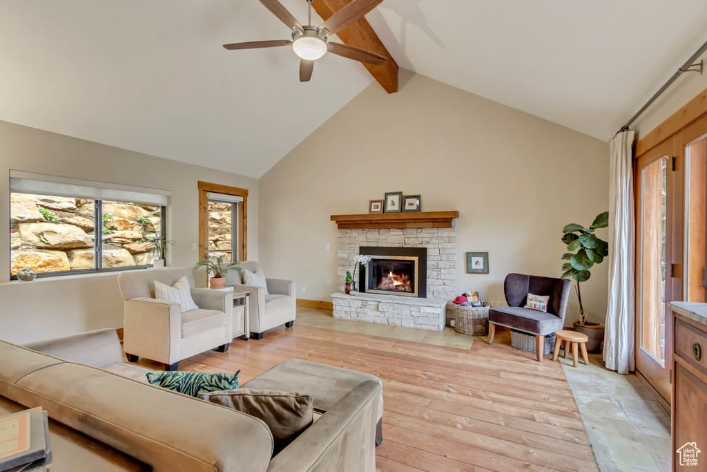 Living room featuring light wood-style flooring, ceiling fan, beam ceiling, a stone fireplace, and high vaulted ceiling