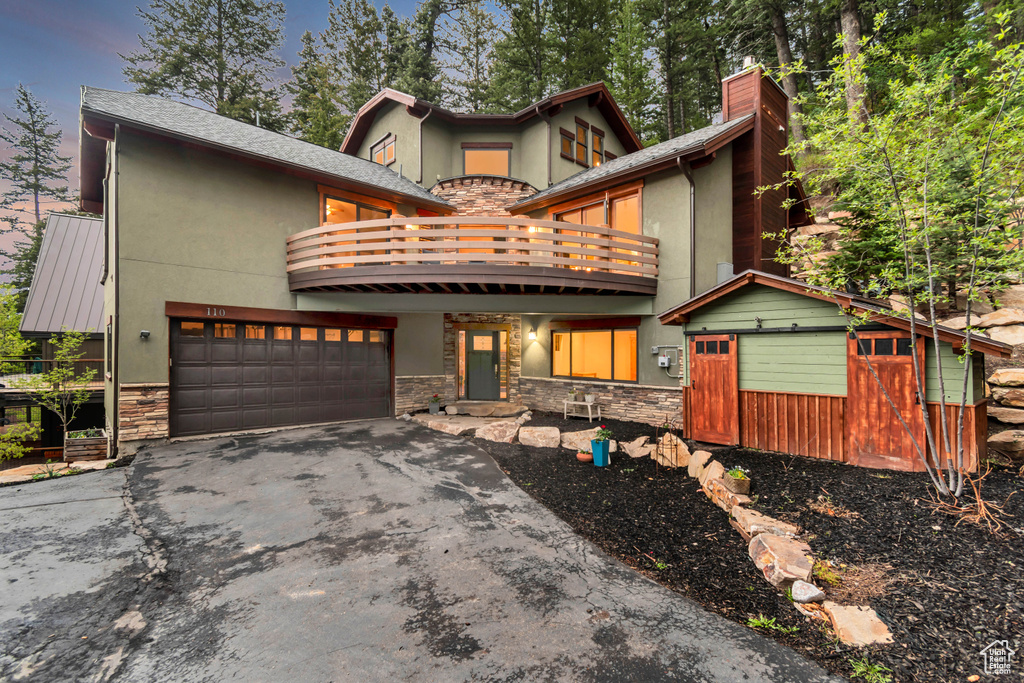 View of front of home with a balcony, stone siding, stucco siding, and driveway