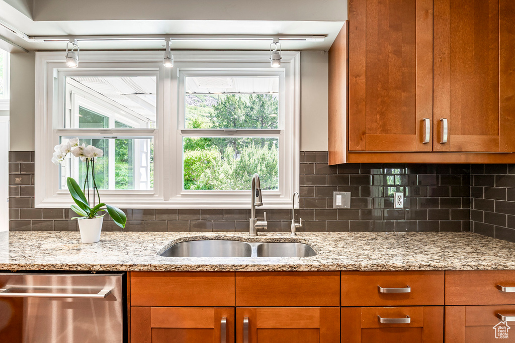 Kitchen with stainless steel dishwasher, a sink, track lighting, light stone counters, and backsplash