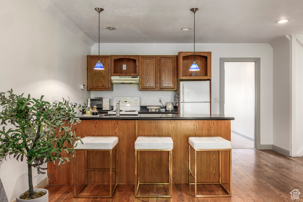Kitchen with white appliances, under cabinet range hood, dark countertops, dark wood-type flooring, and brown cabinetry