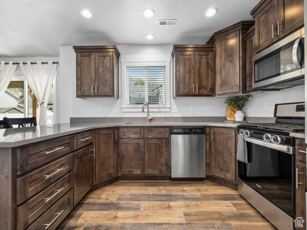Kitchen with appliances with stainless steel finishes, a sink, wood finished floors, a peninsula, and dark brown cabinets