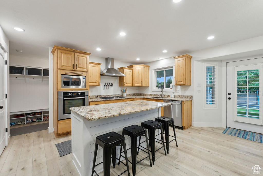 Kitchen featuring stainless steel appliances, wall chimney exhaust hood, light brown cabinetry, light wood finished floors, and a center island