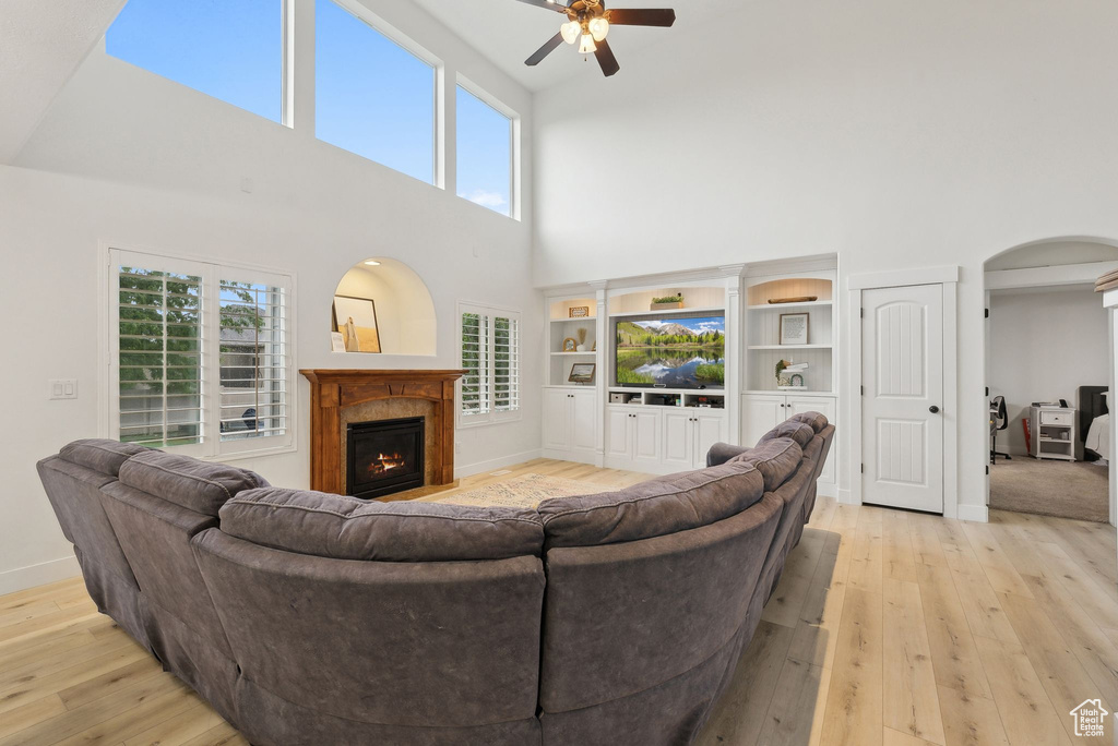 Living room with a towering ceiling, ceiling fan, light wood finished floors, arched walkways, and a warm lit fireplace