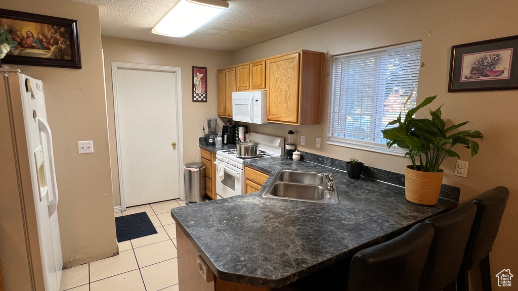 Kitchen featuring white appliances, dark countertops, a peninsula, light tile patterned flooring, and a sink