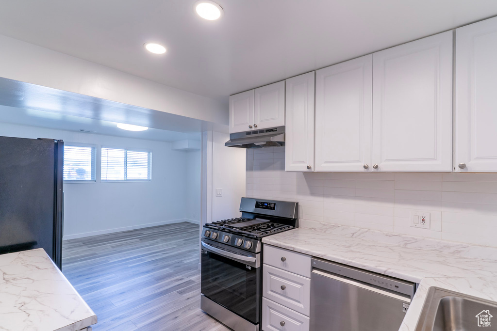 Kitchen with stainless steel appliances, under cabinet range hood, white cabinetry, light wood-type flooring, and backsplash