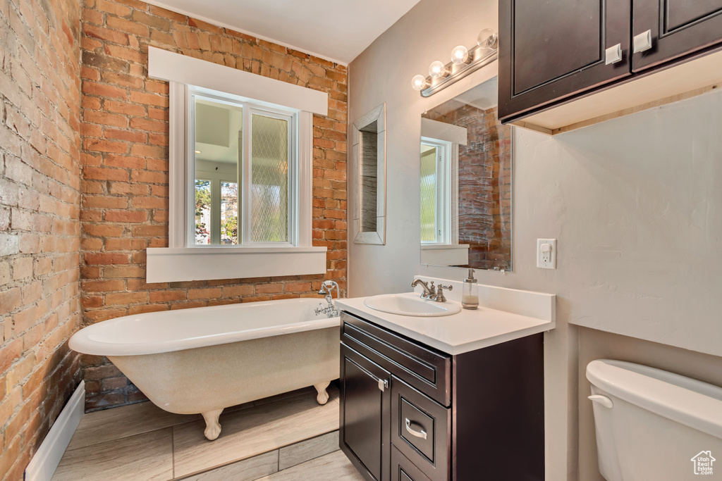 Full bath featuring toilet, a soaking tub, vanity, wood finished floors, and brick wall