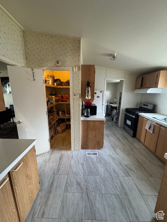 Kitchen with gas range oven, wallpapered walls, brown cabinetry, light countertops, and freestanding refrigerator
