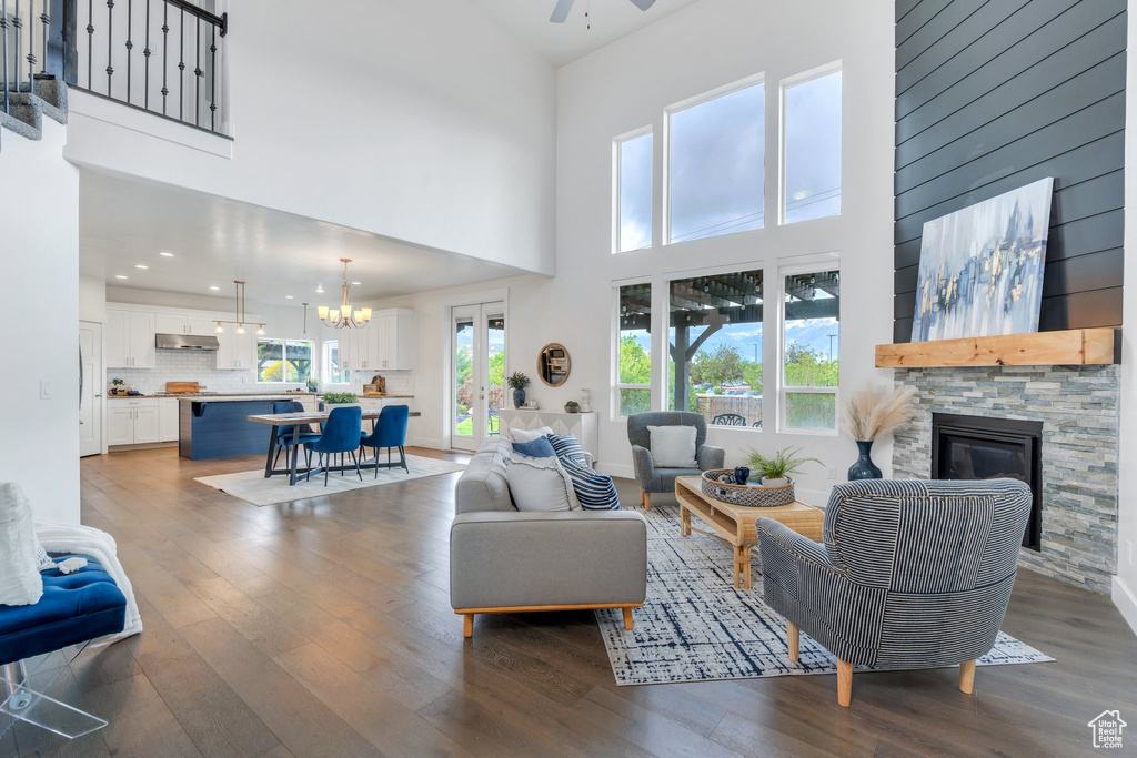 Living room featuring wood finished floors, a stone fireplace, ceiling fan, a towering ceiling, and a chandelier