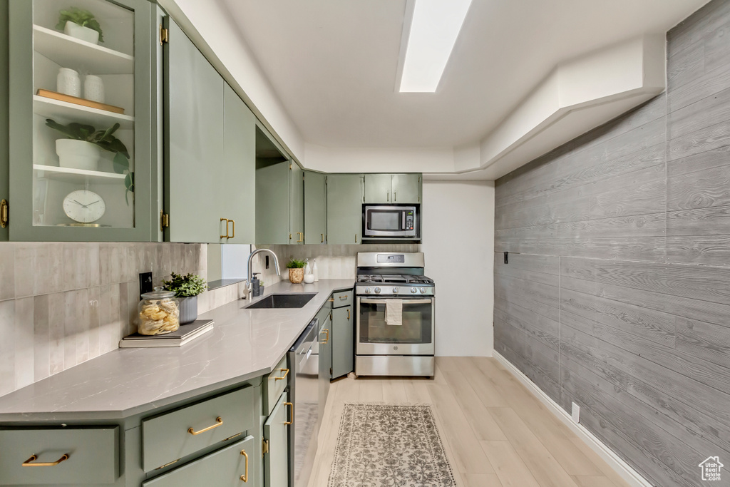 Kitchen with green cabinetry, appliances with stainless steel finishes, a sink, wooden walls, and light wood-style flooring