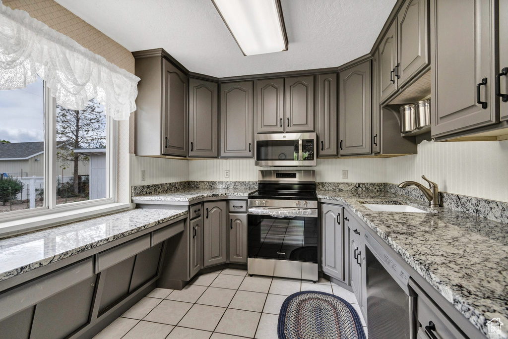 Kitchen featuring appliances with stainless steel finishes, a sink, light tile patterned flooring, gray cabinets, and light stone counters