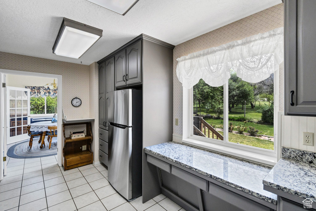 Kitchen featuring freestanding refrigerator, gray cabinetry, light stone counters, light tile patterned floors, and wallpapered walls