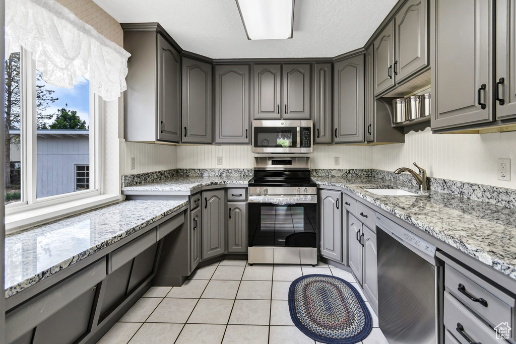Kitchen with appliances with stainless steel finishes, a sink, gray cabinetry, light tile patterned floors, and light stone countertops