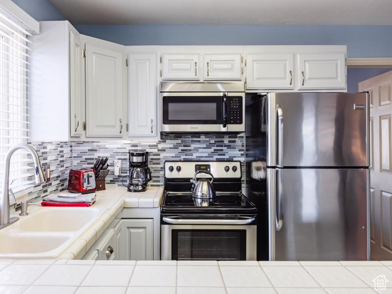 Kitchen with appliances with stainless steel finishes, tile countertops, a sink, white cabinets, and tasteful backsplash