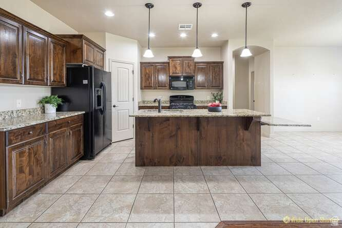 Kitchen featuring black appliances, light tile patterned floors, light stone counters, hanging light fixtures, and a kitchen island with sink