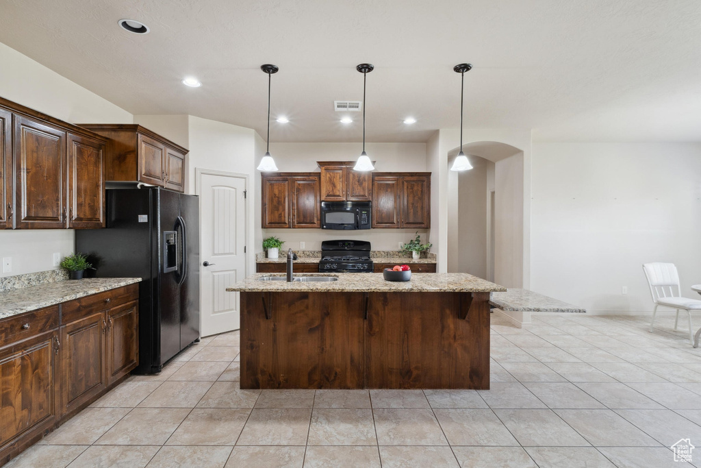 Kitchen with black appliances, a sink, arched walkways, a center island with sink, and light stone countertops