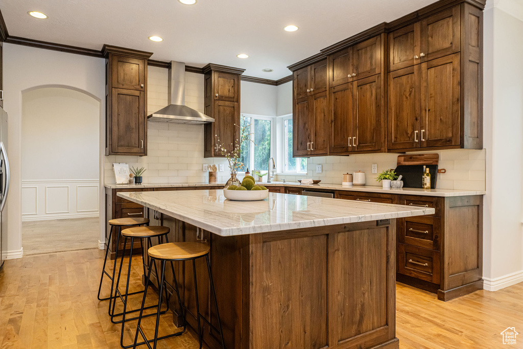 Kitchen featuring wall chimney exhaust hood, crown molding, recessed lighting, light wood-style floors, and a kitchen bar