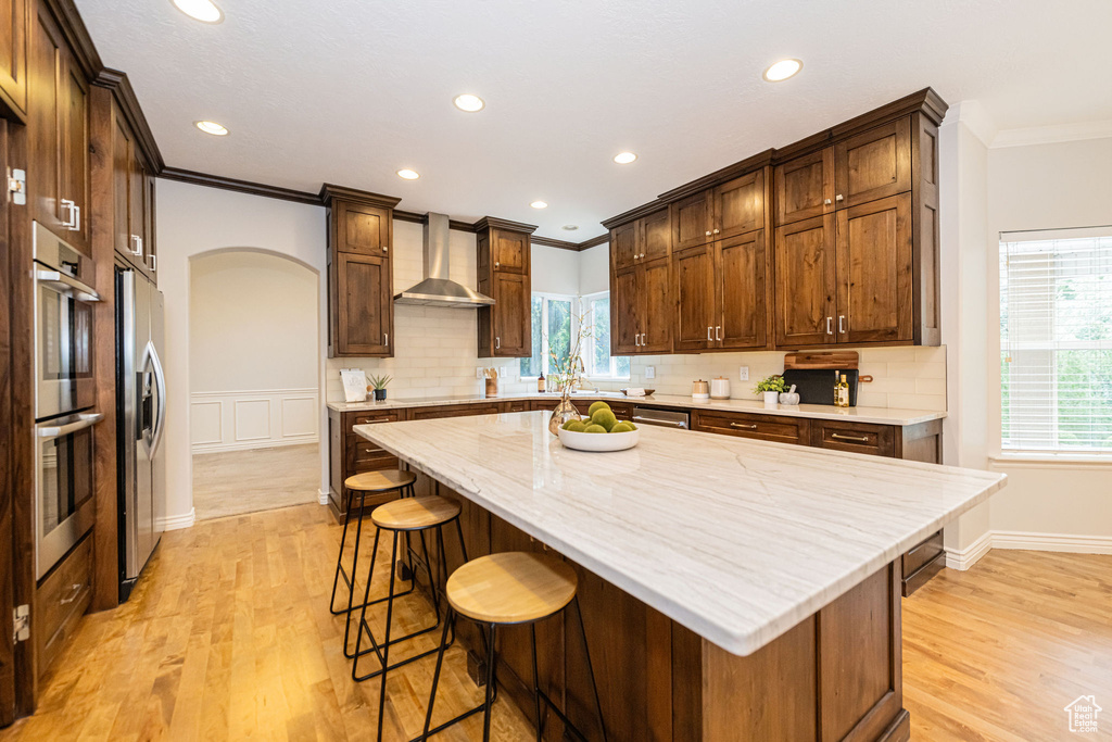 Kitchen with wall chimney exhaust hood, crown molding, arched walkways, stainless steel appliances, and light wood-style flooring