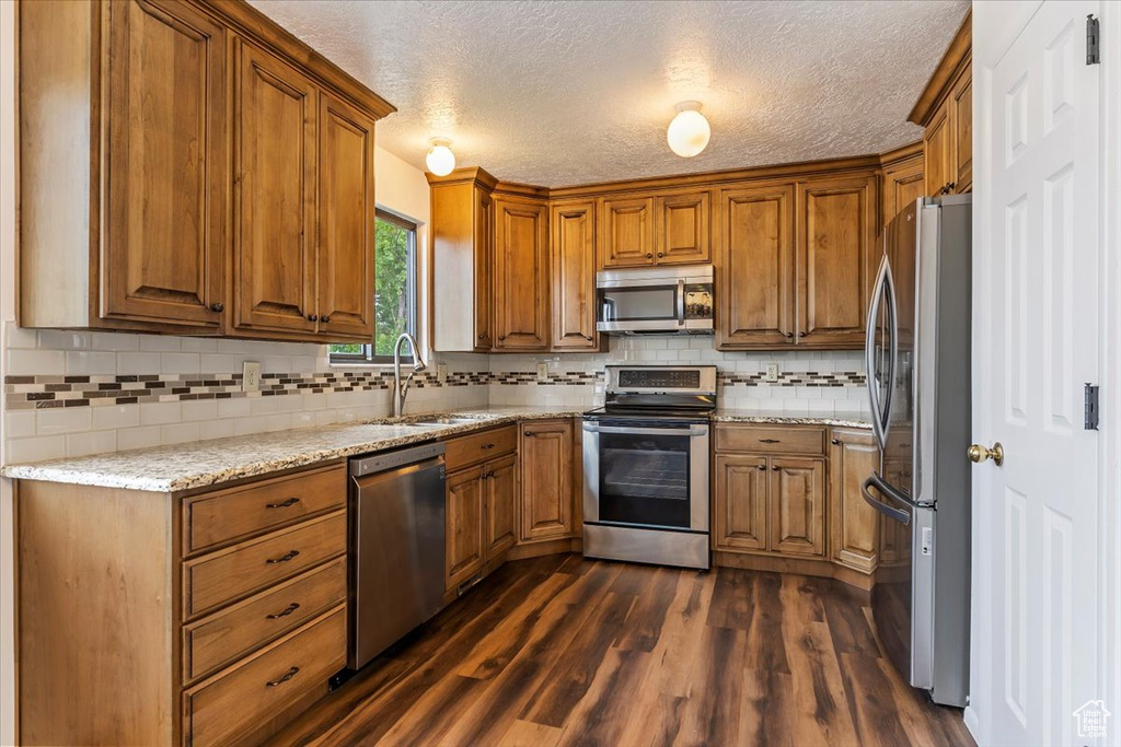 Kitchen with appliances with stainless steel finishes, a sink, brown cabinetry, dark wood-type flooring, and backsplash