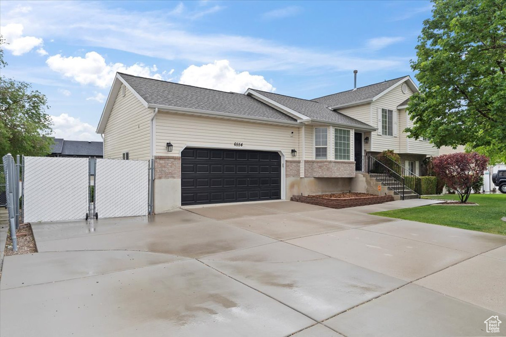 Split level home featuring a garage, driveway, brick siding, roof with shingles, and a gate