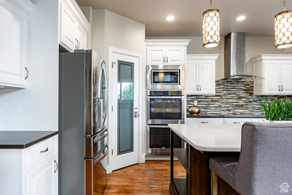 Kitchen featuring appliances with stainless steel finishes, wall chimney range hood, white cabinets, tasteful backsplash, and wood finished floors