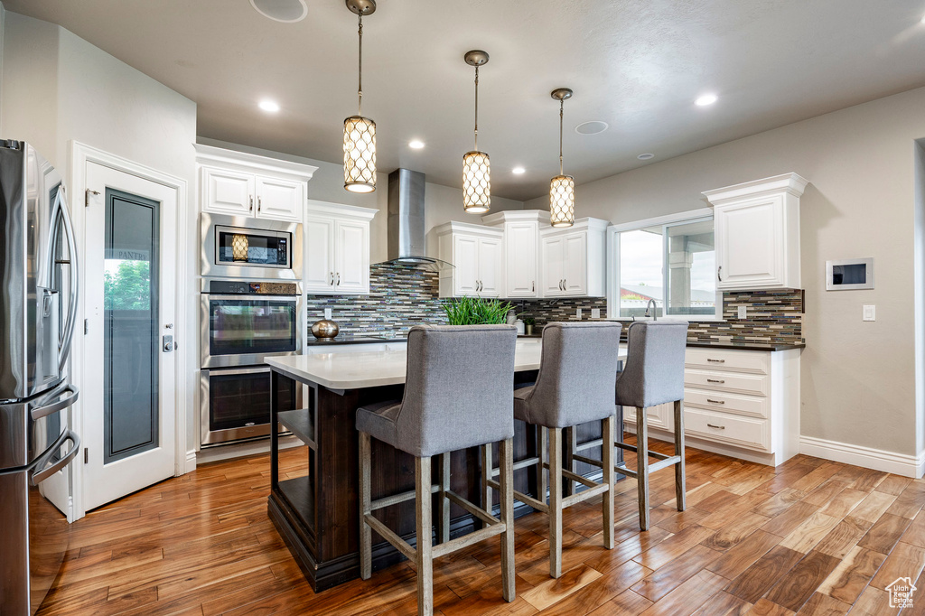 Kitchen featuring wall chimney range hood, stainless steel appliances, tasteful backsplash, light wood-style floors, and white cabinetry