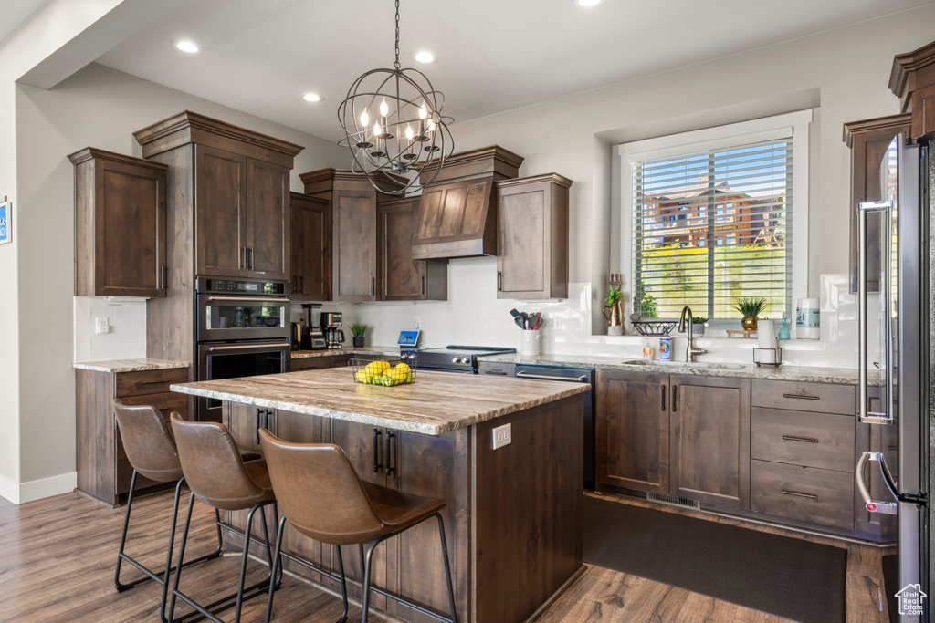 Kitchen with appliances with stainless steel finishes, premium range hood, a center island, wood finished floors, and light stone counters