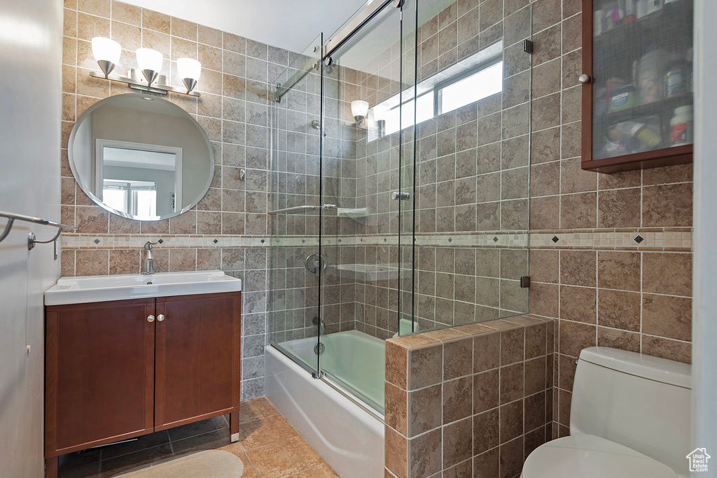 Full bathroom with tile walls, tile patterned flooring, bath / shower combo with glass door, toilet, and vanity