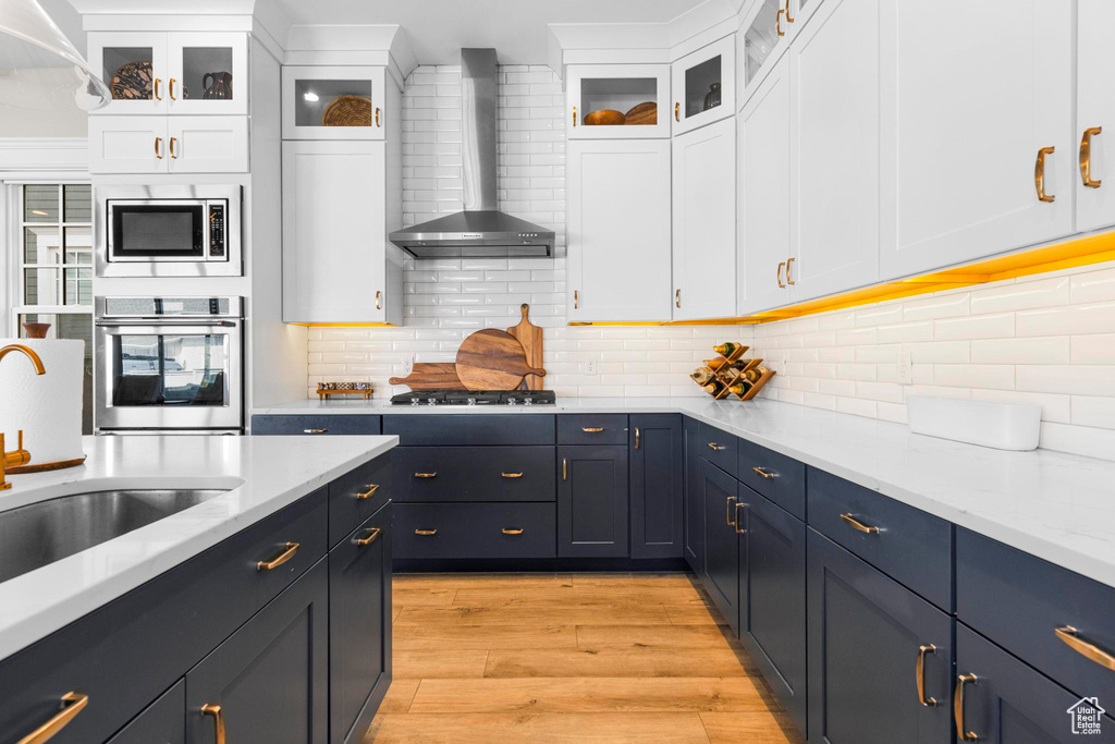 Kitchen featuring appliances with stainless steel finishes, wall chimney exhaust hood, light wood finished floors, white cabinets, and decorative backsplash