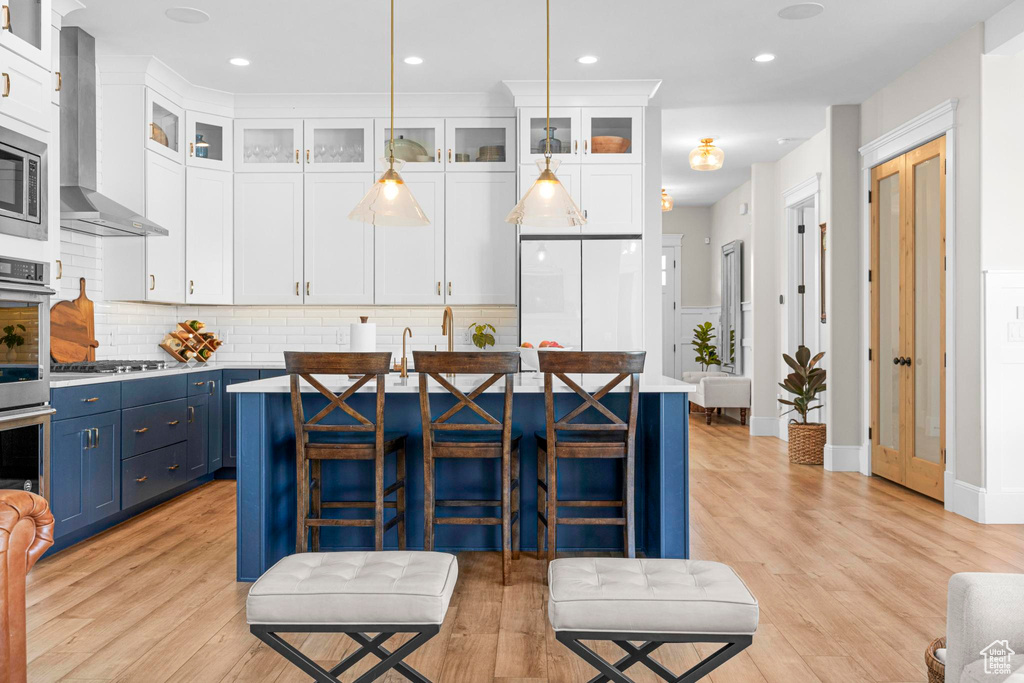 Kitchen with blue cabinets, wall chimney exhaust hood, an island with sink, white cabinetry, and decorative backsplash
