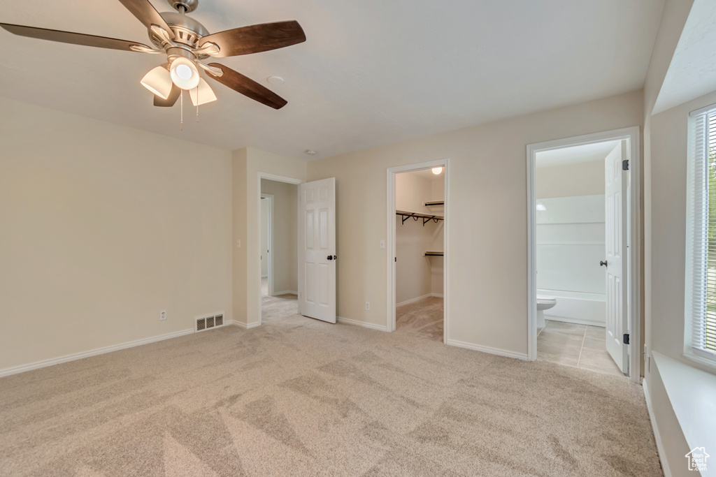 Unfurnished bedroom with light carpet, a spacious closet, ensuite bath, baseboards, and a ceiling fan