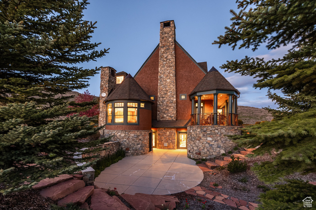 View of front of property featuring a chimney, a balcony, stone siding, a patio area, and a shingled roof