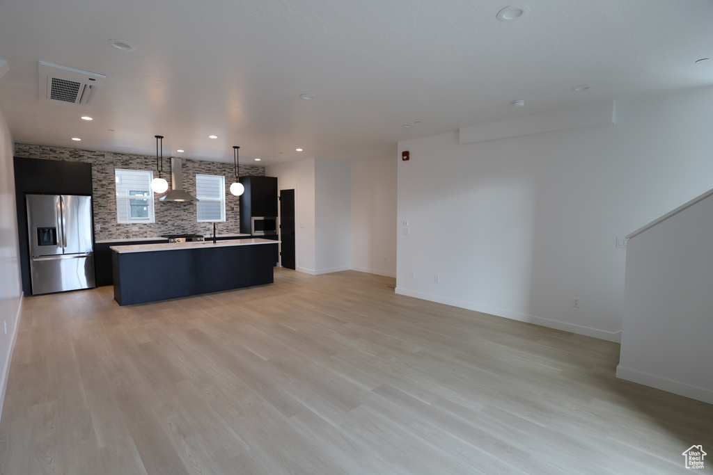 Kitchen featuring stainless steel fridge with ice dispenser, wall chimney range hood, a kitchen island with sink, decorative backsplash, and light countertops