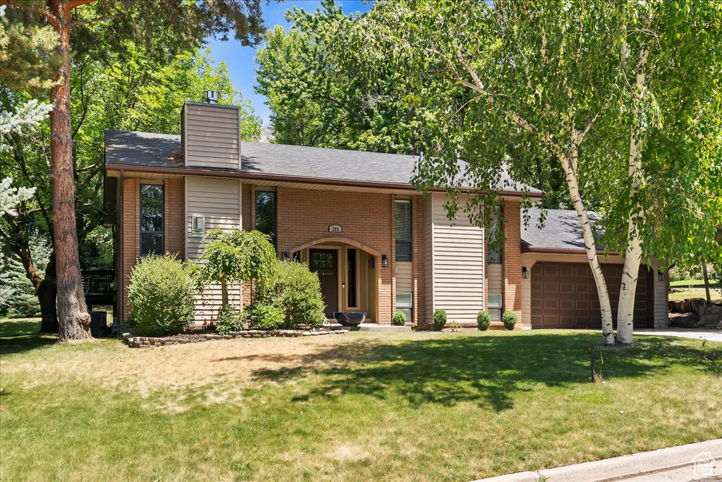 View of front of home with an attached garage, a front lawn, a chimney, brick siding, and a shingled roof