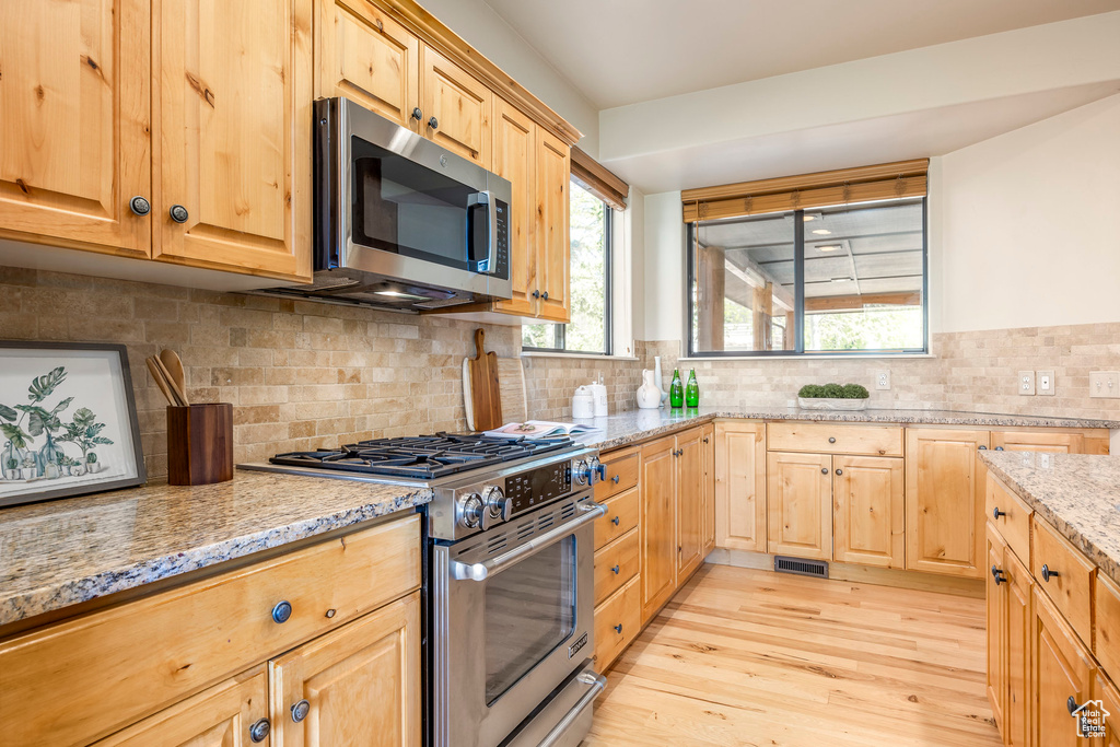 Kitchen with stainless steel appliances, light brown cabinetry, light wood-style flooring, light stone countertops, and decorative backsplash