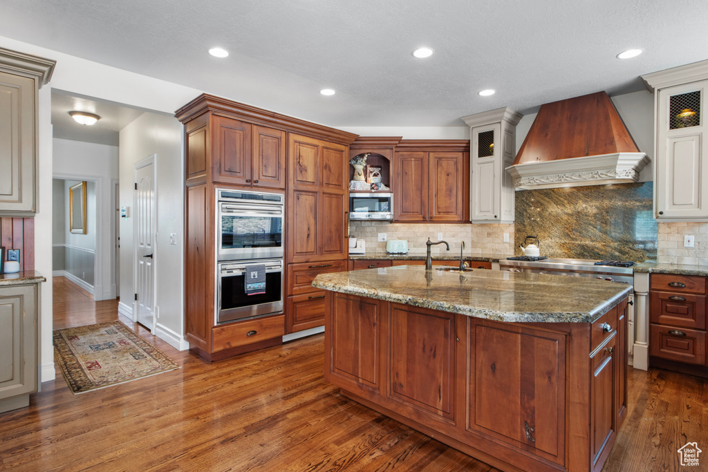 Kitchen with decorative backsplash, appliances with stainless steel finishes, custom range hood, and glass insert cabinets