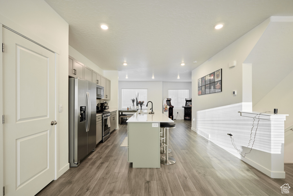 Kitchen with appliances with stainless steel finishes, a kitchen bar, wood finished floors, a kitchen island with sink, and light countertops