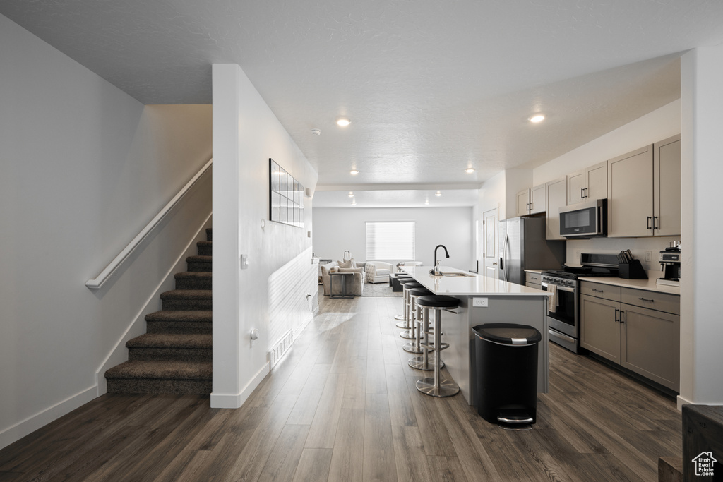 Kitchen featuring stainless steel appliances, a sink, gray cabinetry, a center island with sink, and dark wood-type flooring