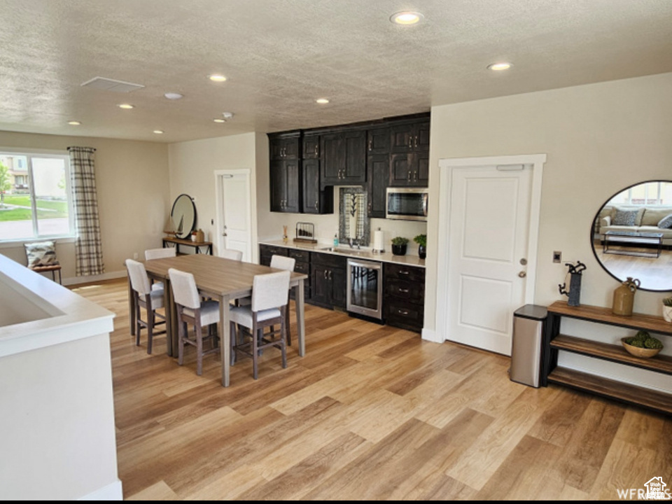 Kitchen featuring wine cooler, stainless steel microwave, dark cabinets, recessed lighting, and light wood-type flooring
