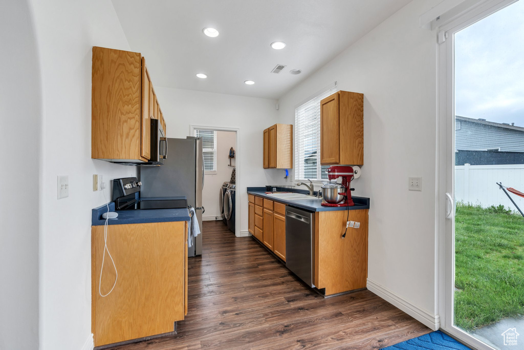 Kitchen with appliances with stainless steel finishes, a sink, washing machine and clothes dryer, dark wood-style floors, and dark countertops