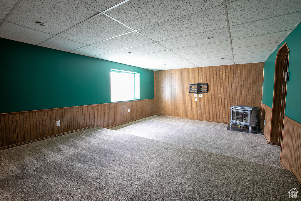 Carpeted empty room featuring a wood stove, wood walls, a paneled ceiling, and wainscoting