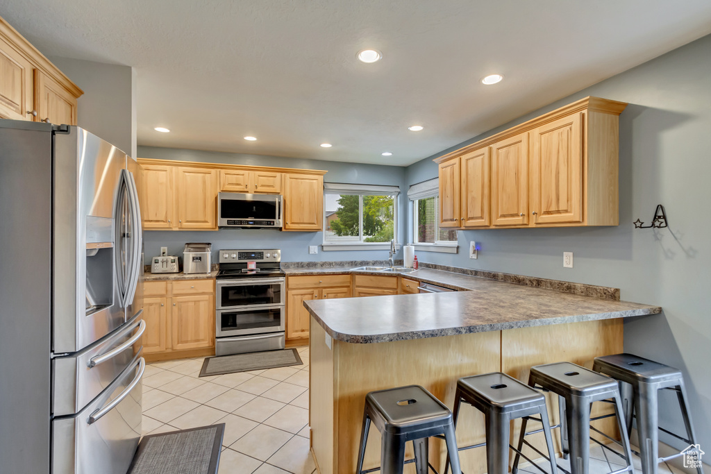 Kitchen with stainless steel appliances, a sink, a peninsula, light brown cabinets, and a kitchen bar