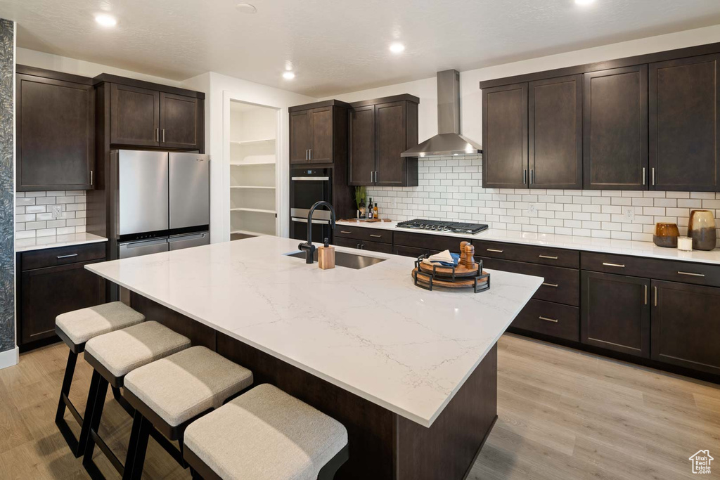 Kitchen with wall chimney range hood, a sink, appliances with stainless steel finishes, a kitchen breakfast bar, and light wood-style flooring