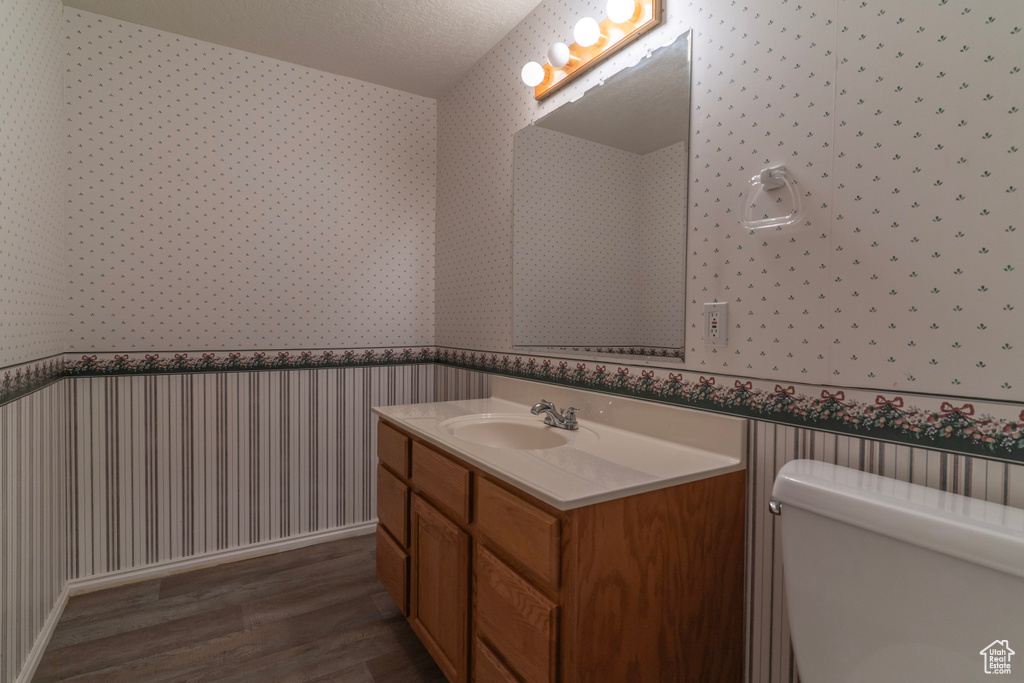 Bathroom with wallpapered walls, wood finished floors, a wainscoted wall, vanity, and toilet