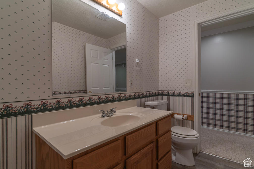 Bathroom featuring wallpapered walls, toilet, wood finished floors, vanity, and a wainscoted wall