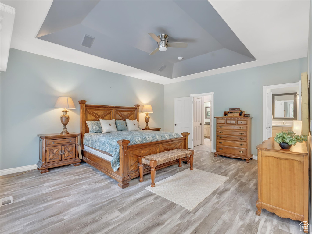 Bedroom with a tray ceiling, baseboards, light wood-type flooring, connected bathroom, and ceiling fan