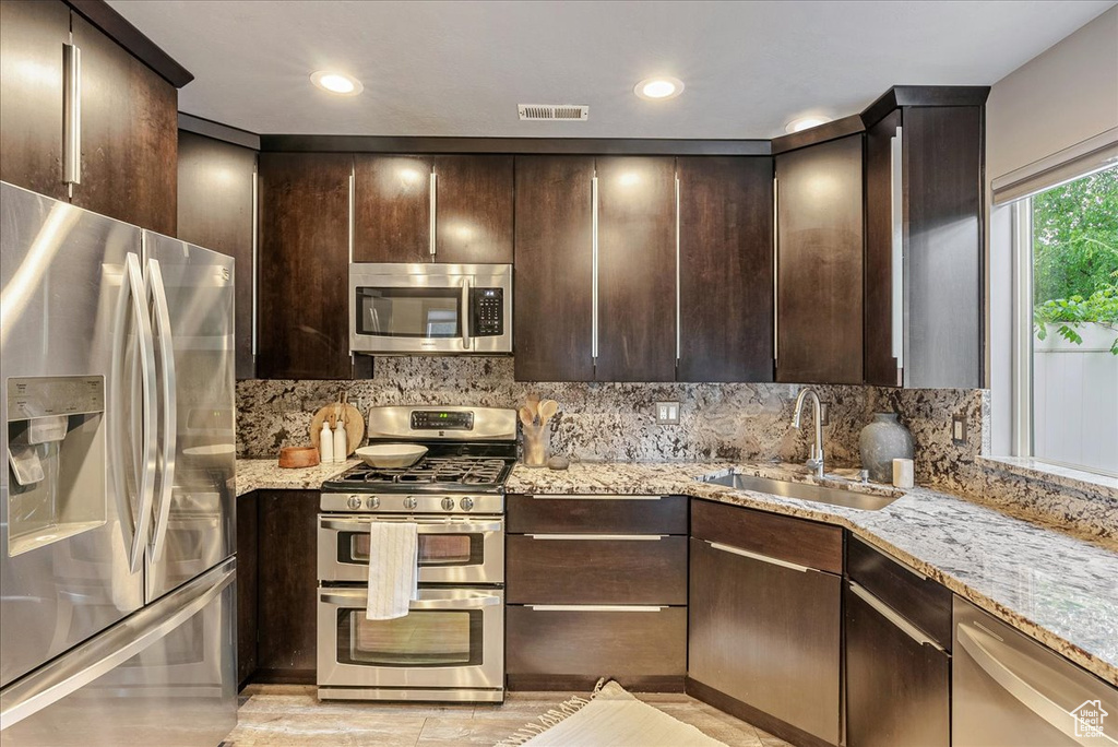 Kitchen featuring appliances with stainless steel finishes, a sink, dark brown cabinetry, light stone counters, and recessed lighting
