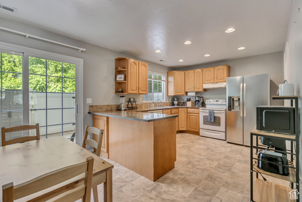 Kitchen with white electric range oven, stainless steel refrigerator with ice dispenser, a peninsula, open shelves, and light brown cabinets