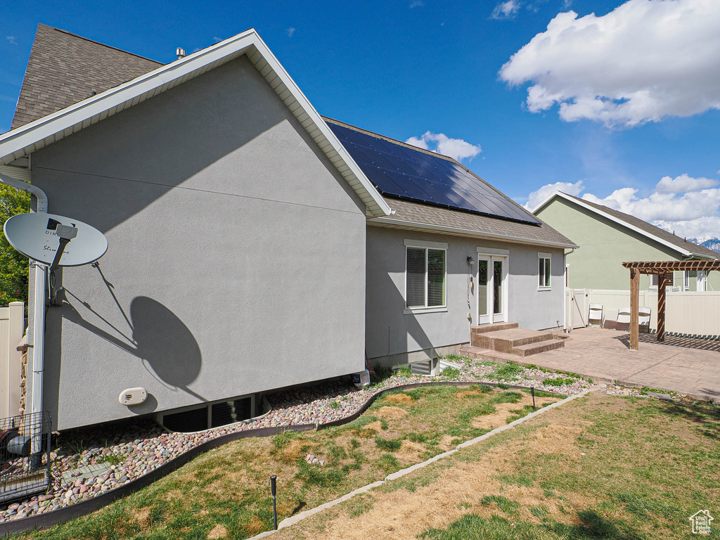 Rear view of property featuring solar panels, stucco siding, a pergola, a shingled roof, and a patio area